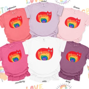 May include: A collection of six t-shirts in various colors, including watermelon, orchid, blossom, crunchberry, white, and berry. Each shirt features a red cat design with a rainbow-colored cat inside. The shirts are casual wear.