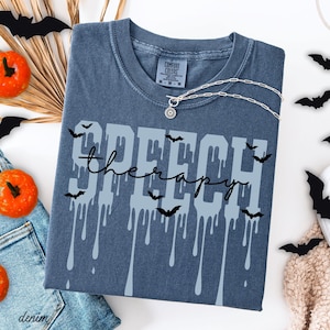 May include: A blue t-shirt with the words "SPEECH therapy" in a dripping font, decorated with bat silhouettes. A silver necklace with a sun pendant is on the shirt. The shirt is surrounded by Halloween decorations, including pumpkins and paper bats.