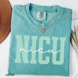 May include: Seafoam green t-shirt with the word "NICU" in large, light green block letters and "nicu" in a cursive font. A gold necklace with a small pendant rests on the shirt. The shirt is labeled "Comfort Colors".