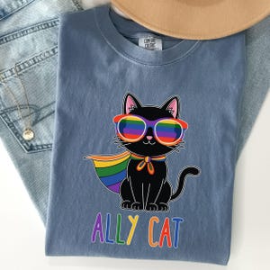 May include: A blue Comfort Colors t-shirt featuring a black cat wearing rainbow sunglasses and a rainbow cape. The text "ALLY CAT" is printed in rainbow colors below the cat. The shirt is folded on top of blue jeans and a tan hat.