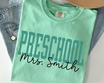 Może przedstawiać: Jasnozielona koszulka z napisem "PRESCHOOL" w turkusowych blokowych literach i "Mrs. Smith" w czarnym kursywie. Koszulka jest złożona na niebieskich dżinsach i brązowym kapeluszu.