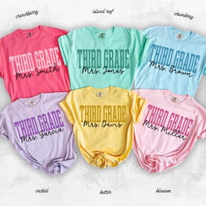 May include: Six colorful Third Grade t-shirts in shades of pink, blue, green, yellow, and purple. Each shirt features the words "THIRD GRADE" in bold letters, with a teacher's name below. The shirts are labeled with their color names.