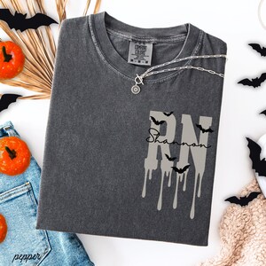 May include: A charcoal gray short-sleeve t-shirt with a Comfort Colors label at the neck. The front features a large, light gray, dripping-style 'RN' graphic with small black bat silhouettes and the name 'Shannon' written in black script.