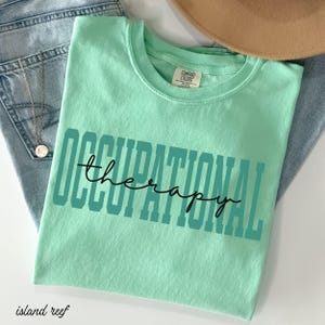 May include: A light green t-shirt with the words "OCCUPATIONAL THERAPY" printed in teal and black. The shirt is folded and placed on top of blue jeans and a gold necklace.