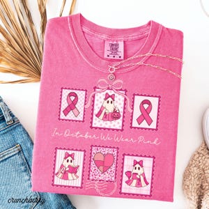 May include: Pink Comfort Colors t-shirt with a graphic design featuring pink ribbons and a ghost character. The shirt has the text "In October We Wear Pink". A silver necklace is draped across the shirt.
