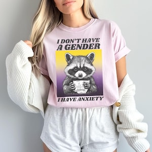 May include: A pink t-shirt with a black and white illustration of a raccoon holding a coffee cup. The text on the shirt reads "I don't have a gender I have anxiety".