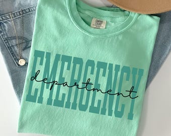 May include: A light green t-shirt with the words "EMERGENCY department" printed in teal and black. The shirt is folded on top of blue jeans, with a gold necklace and a tan hat in the background.