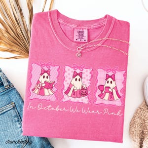 May include: A pink Comfort Colors t-shirt with the text "In October We Wear Pink." The shirt features three ghost illustrations with pink bows and ribbons. A silver necklace with a flower pendant is on the shirt.