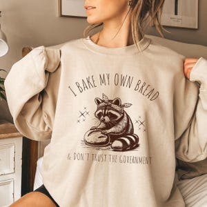 May include: A beige sweatshirt with a brown raccoon graphic and the text "I bake my own bread & don't trust the government".