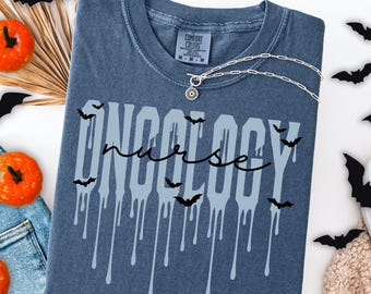 May include: A denim blue t-shirt with the words "ONCOLOGY nurse" in a dripping font, decorated with bat silhouettes. A silver necklace with a sun pendant is on the shirt. The shirt is part of a Halloween-themed collection.