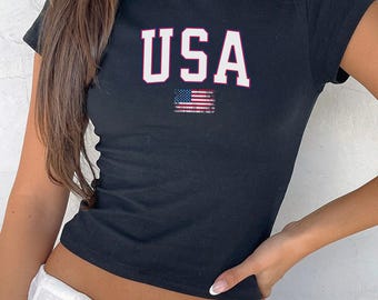 May include: Black crop top with the word "USA" in white block letters with a red outline. Below the text is a distressed American flag graphic. The shirt has short sleeves and a scoop neckline.