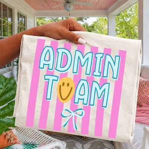 May include: An ivory-colored t-shirt featuring vertical pink stripes and the text ADMIN TEAM in light blue letters. A yellow smiley face replaces the letter I, and a light blue bow graphic is positioned below the text on the front.