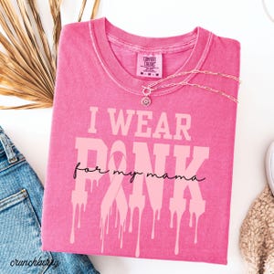 May include: A pink Comfort Colors t-shirt with the text "I WEAR PINK for my mama" in a light pink font. The word "PINK" has a dripping effect. A silver necklace with a floral pendant is on the shirt.