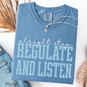 Pode incluir: Uma camiseta azul Comfort Colors com as palavras "alright stop REGULATE AND LISTEN" em branco e azul claro. Um colar de prata com um pingente de sol está drapeado sobre a camisa. A camisa está dobrada sobre uma superfície branca.