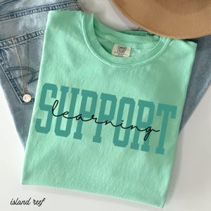 May include: A light green t-shirt with the words "SUPPORT learning" printed in teal block letters and black cursive. The shirt is folded on top of blue jeans and a gold necklace.