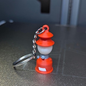 Puede incluir: Un llavero de linterna roja y blanca en miniatura. La linterna tiene un diseño detallado con una cadena y un llavero plateados. La linterna mide aproximadamente 4 cm de altura.