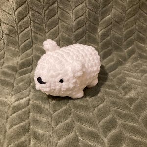 May include: A small, white, crocheted bunny plush toy with black bead eyes and a black nose. The bunny is sitting on a green, textured blanket. The plush toy has a rounded body and small ears.