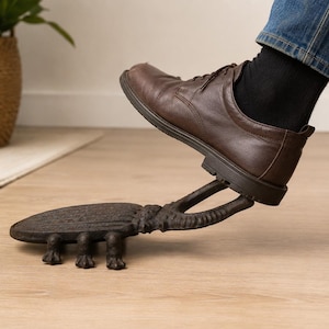 May include: A dark brown cast iron boot jack shaped like a beetle with a textured shell and six legs. The device features a U-shaped opening at the head to grip the heel of a shoe, allowing for hands-free removal of footwear while standing.