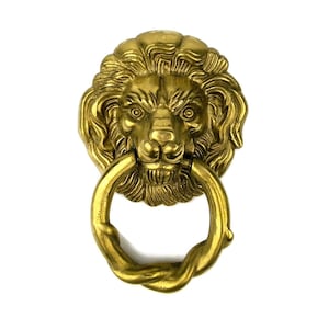 Puede incluir: Un llamador de puerta de cabeza de león dorado con un asa de anillo.