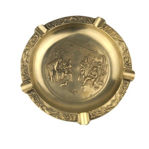 May include: A gold-colored metal ashtray with a raised design of people in a scene. The ashtray has four indentations around the edge.
