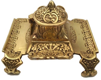 May include: A gold-toned metal inkwell with an ornate design. The inkwell has a hinged lid and sits on a decorative base with intricate details.