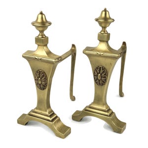 May include: Pair of antique brass andirons. Each andiron features a decorative floral medallion, a curved handle, and a finial. The andirons are made of brass and have a gold finish. They are designed to hold logs in a fireplace.