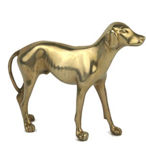 May include: A brass dog figurine in a standing pose. The sculpture has a smooth, polished surface with a golden hue. The dog's features are detailed, including its ears, snout, and tail. The figurine is a decorative item.