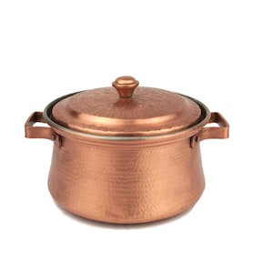 Puede incluir: Una olla de cocina de color cobre con tapa y dos asas. La olla tiene una textura martillada y una forma redondeada. La tapa tiene un pequeño pomo en la parte superior. La olla está diseñada para cocinar y servir alimentos.