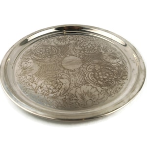 May include: A circular, silver-toned serving tray featuring an elaborate embossed floral and scroll pattern. The tray has a gently raised rim and a polished surface, ideal for serving food or displaying decorative objects.