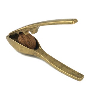 May include: A vintage brass nutcracker with a walnut inside. The nutcracker is open, ready to crack the nut. The brass has a warm, aged appearance. The background is plain white.
