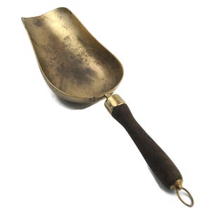 May include: A brass scoop with a wooden handle. The scoop is curved and has a worn, aged look. The handle is dark brown and has a brass ring at the end.