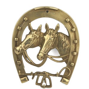 Könnte beinhalten: Messing Wandbehang in Form eines Hufeisens, das zwei Pferdeköpfe darstellt. Die Pferde sind mit Zäumen detailliert und befinden sich innerhalb des Hufeisens. Ein dekoratives Gebiss und Zügel befinden sich darunter.
