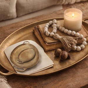 Könnte beinhalten: Ein goldfarbenes ovales Tablett mit Griffen enthält eine brennende Kerze, einen Stapel Bücher, dekorative Perlen und zwei kleine Tabletts. Zwei Walnüsse und eine Quaste ergänzen die Dekoration. Die Gegenstände liegen auf einer Holzoberfläche.