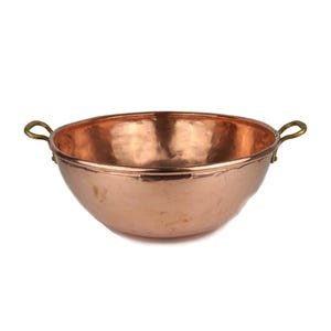 May include: A copper bowl with a rounded shape and two brass handles. The bowl has a reflective surface and a slightly hammered texture. The bowl is likely used for cooking or serving.