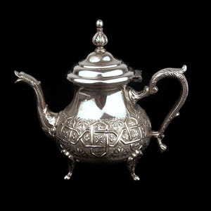 Peut inclure: Une théière argentée avec des motifs floraux et géométriques complexes. La théière a un bec verseur incurvé, une anse décorative et un couvercle avec un fleuron. Elle repose sur trois petits pieds sur fond noir.