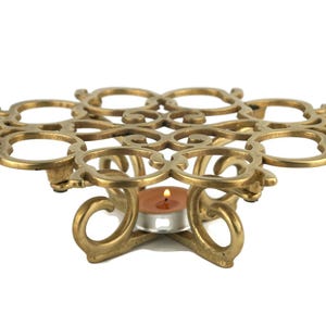 Può includere: Un portacandele in metallo dorato con un design decorativo e ornato. Il portacandele ha una parte superiore rotonda e una base con tre gambe. Una piccola candela da tè è posizionata al centro del portacandele.