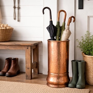 May include: A rustic entryway scene featuring a wooden bench with a woven basket and brown leather boots. A copper umbrella stand holds several umbrellas. A woven basket with greenery and green rain boots complete the decor.