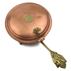 May include: A copper-coloured, round serving dish with a hinged handle and decorative brass accents. The dish has a small brass emblem on the lid and rests on three small feet. The handle features a leaf design.