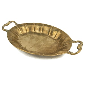 May include: Oval-shaped brass tray with two ornate handles. The tray has a fluted design around the edges and a slightly aged appearance. The brass color is a warm, golden hue, and the tray is set against a white background.
