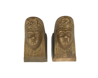 Puede incluir: Un par de sujetalibros de metal dorado con un rostro femenino estilizado con una corona. Los sujetalibros tienen la forma de un bloque rectangular con un diseño en relieve.