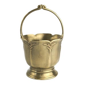 May include: A small, antique-style brass bucket with a handle. The bucket has a scalloped rim and decorative leaf-like patterns. The handle is arched and attached to the bucket with decorative flourishes. The bucket sits on a small, round base.