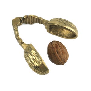 May include: A vintage brass nutcracker with a walnut-shaped design. The nutcracker is open, revealing the cracking mechanism, and a single walnut sits beside it. The brass has a warm, golden hue.