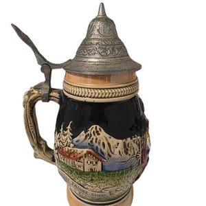 Peut inclure: Une chope à bière en céramique avec un couvercle métallique et un levier à pouce articulé. Le corps présente un relief représentant un paysage de montagne, une maison et des pins sur fond noir, avec le texte allemand "LEBE TRINKE SINGE".