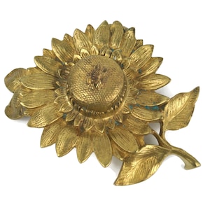 Op de afbeelding: Een gouden decoratief object in de vorm van een zonnebloem. Het object heeft gedetailleerde bloemblaadjes, een getextureerd midden en een stengel met bladeren. De zonnebloem is gemaakt van metaal en heeft een vintage uitstraling. Het object is waarschijnlijk een decoratief stuk of een broche.