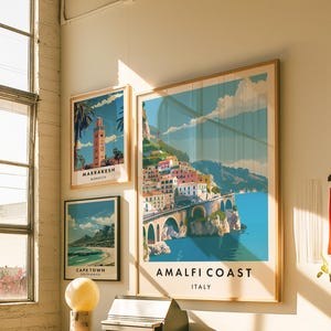 May include: Three framed travel posters on a white wall. The posters feature colorful illustrations of Amalfi Coast, Italy, Marrakesh, Morocco, and Cape Town, South Africa.