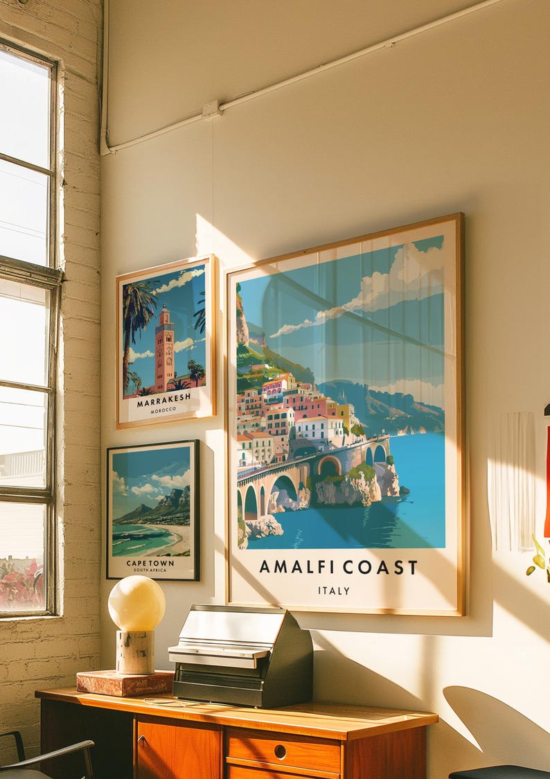 May include: Three framed travel posters on a white wall. The posters feature colorful illustrations of Amalfi Coast, Italy, Marrakesh, Morocco, and Cape Town, South Africa.