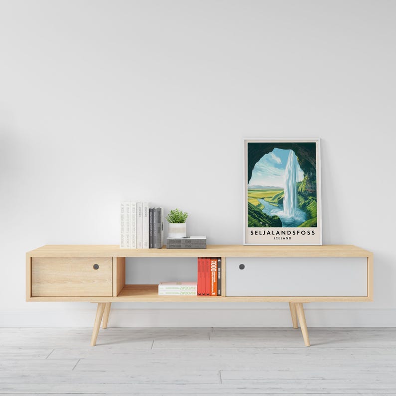 May include: A framed print of Seljalandsfoss waterfall in Iceland, displayed on a light wood console table. The print features a waterfall cascading into a river, with green hills and a blue sky. The table has a drawer and a cabinet.