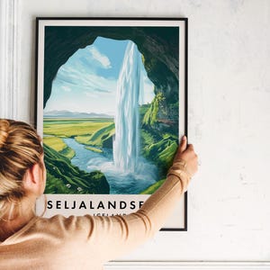May include: A framed print featuring a scenic waterfall landscape. The artwork depicts a waterfall cascading into a river, framed by a cave opening. The print includes the text "SELJALANDSFOSS ICELAND" at the bottom. The print is held by a person.