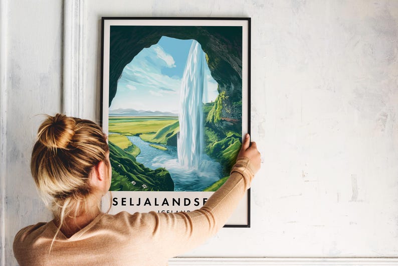 May include: A framed print featuring a scenic waterfall landscape. The artwork depicts a waterfall cascading into a river, framed by a cave opening. The print includes the text "SELJALANDSFOSS ICELAND" at the bottom. The print is held by a person.