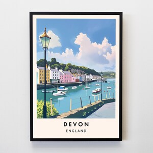 May include: A framed print of Devon, England, showing a coastal town. The artwork features colorful buildings along a blue waterway with boats, under a bright blue sky with fluffy clouds. The print is in a black frame.